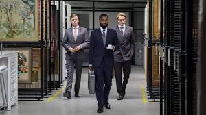 Three men in suits walking down an art-lined corridor, one holding a briefcase and coffee, from Tenet.